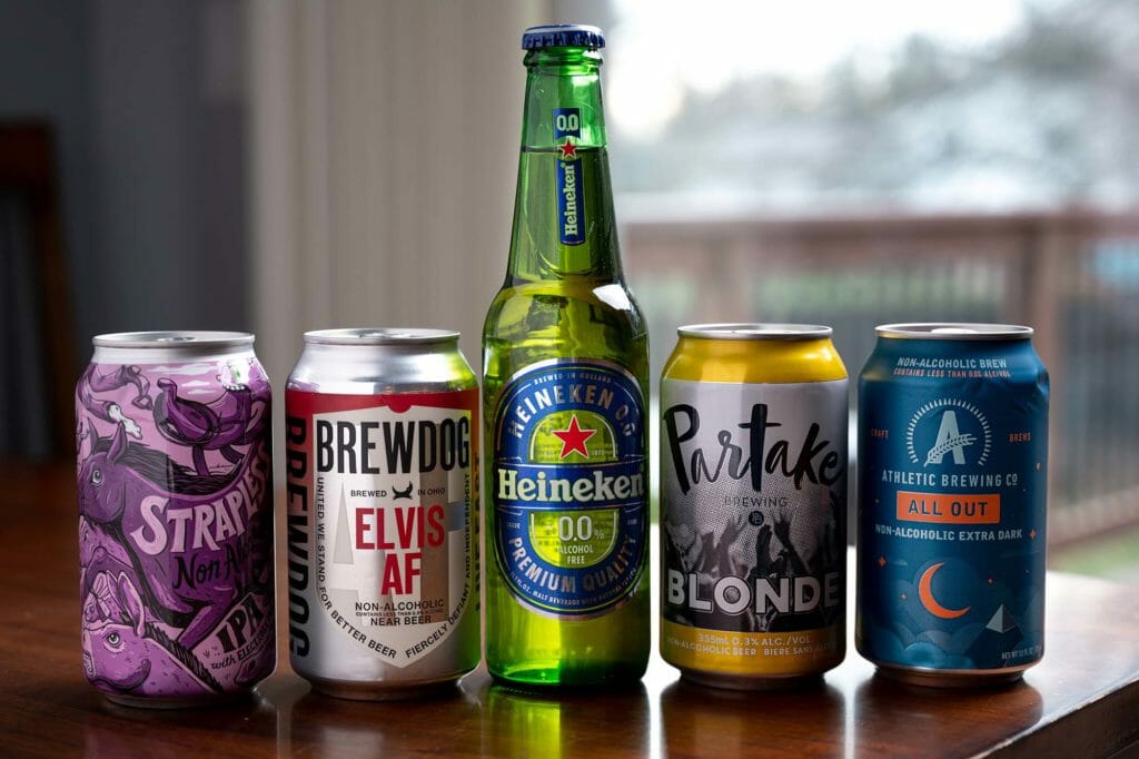 Quatro latas e uma garrafa de cerveja sem álcool estão alinhadas sobre uma mesa.